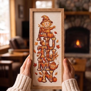 Könnte beinhalten: Eine vertikal gerahmte Kreuzstichstickerei mit dem Wort AUTUMN in großen, karierten Buchstaben. Eine lächelnde Vogelscheuche sitzt auf den Buchstaben, umgeben von kleinen orangefarbenen Kürbissen, einer Teekanne und verstreuten Herbstblättern.