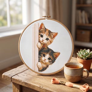 May include: A circular wooden embroidery hoop containing a cross-stitch pattern of two kittens peeking from behind a vertical edge. The top kitten is orange and white, while the bottom kitten is dark brown, black, and white, both with large eyes.