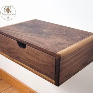 May include: A rectangular floating wall shelf made of dark brown walnut wood with a single drawer. The drawer front features a semi-circular cutout for opening. The wood grain is visible across the top and front surfaces of the unit.