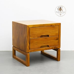 May include: A light brown wooden nightstand featuring two drawers with integrated horizontal handle cutouts. The piece is supported by a rectangular frame base and displays a visible wood grain pattern. A circular logo with the text Timber Muse Aura is visible in the upper right corner.