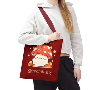 Op de afbeelding: Een donkerrode tote bag met een afbeelding van een schattige paddenstoel met een rode hoed en witte stippen, omringd door kleine oranje blaadjes. De tekst "Shroomtastic" is in een lichtbruin lettertype onderaan het ontwerp gedrukt.