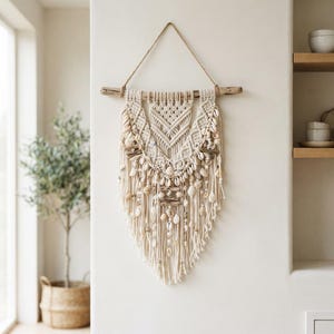 Puede incluir: Un tapiz de pared hecho con cuerda de macramé color hueso anudada en un patrón en forma de V, suspendido de una pieza horizontal de madera flotante. La pieza está decorada con varias conchas pequeñas y cuentas de madera, con flecos largos.