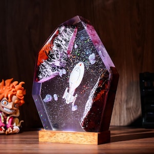 May include: A clear resin sculpture shaped like a jagged crystal, featuring a miniature white starship floating inside among glittery space-like particles. The piece is framed by dark, textured wood elements and rests on a rectangular wooden base.