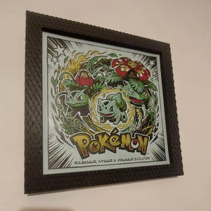 Puede incluir: Un marco negro cuadrado que contiene una impresión de Bulbasaur, Ivysaur y Venusaur en una composición circular. La obra presenta el logotipo de Pokémon y el texto "BULBASAUR, IVYSAUR & VENUSAUR EVOLUTION" en la parte inferior sobre un fondo de cómic.