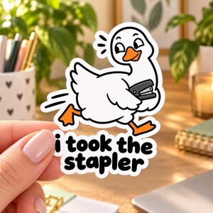 Può includere: Un adesivo in vinile fustellato raffigurante un'oca bianca dei cartoni animati con piedi e becco arancioni, che corre tenendo in mano una cucitrice nera. Sotto l'oca, il testo recita i took the stapler in lettere minuscole nere, arrotondate e in grassetto.