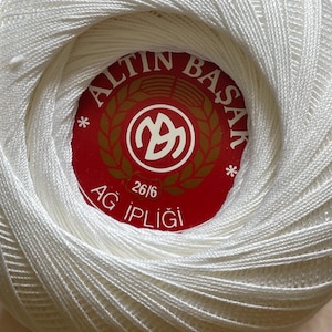 May include: A ball of white crochet thread wrapped in a tight, spherical shape. A circular red label is centered on the top, featuring the white text ALTIN BASAK, the number 26/6, and the words AG IPLIGI, along with a central white logo design.