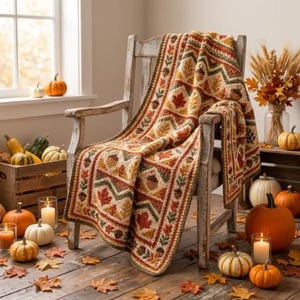 May include: A crochet blanket featuring a repeating pattern of autumn leaves and acorns in shades of orange, brown, yellow, and green. The blanket is draped over a wooden chair, with various pumpkins and dried wheat stalks arranged nearby.