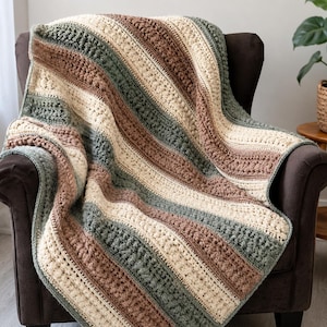 May include: A crochet blanket featuring vertical stripes in alternating shades of cream, sage green, and warm brown. The textile displays a textured pattern with raised bobble stitches and smooth rows, creating a varied surface across the fabric.