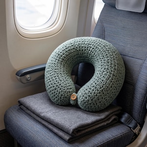 May include: A sage green U-shaped neck pillow made of thick, textured crochet yarn. The pillow features a small, round, light brown wooden button closure at the front centre, designed to secure the two ends of the pillow together.