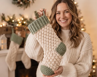 May include: A cream-colored crocheted Christmas stocking featuring a textured bobble stitch pattern, accented with a sage green ribbed cuff, heel, and toe. The stocking includes a small loop at the top for hanging and contains a sprig of pine needles.