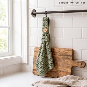 Puede incluir: Una toalla de cocina colgante de ganchillo en color verde salvia con textura de punto de gofre, asegurada con un botón de madera clara en un lazo. El artículo cuelga de una barra de metal oscuro, con el texto CROCHET PATTERN PDF visible.