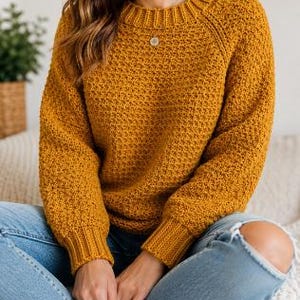 Peut inclure: Un pull à manches longues jaune moutarde avec un motif en tricot texturé, un col rond côtelé et des poignets côtelés, associé à un jean bleu clair avec des détails déchirés aux genoux et un petit collier pendentif circulaire.