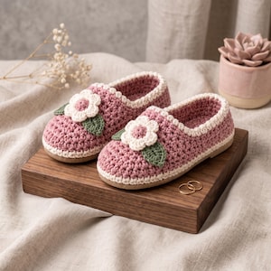 May include: A pair of mauve crochet slippers featuring a white crocheted flower with a pink centre and a green leaf on the toe of each shoe. The slippers have a cream-coloured trim around the opening and a flat, light-coloured sole.