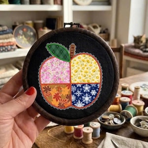 Könnte beinhalten: Ein runder hölzerner Stickrahmen mit schwarzem Stoff, der ein Apfelmotiv in vier Quadranten zeigt. Jeder Quadrant hat ein anderes saisonales Muster: rosa Blumen, gelbe Blüten, Herbstblätter und weiße Schneeflocken, alles mit weißer Spitze umrandet.