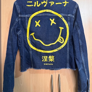 May include: A dark blue denim jacket featuring a large, distressed yellow smiley face graphic on the back. Above the graphic is Japanese text, and below it is the word NIRVANA in English with Japanese characters underneath. The jacket has a raw hem.