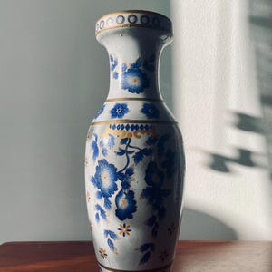 May include: A white ceramic vase featuring a hand-painted floral pattern in shades of blue and gold. The design includes blue flowers, gold leaves, and horizontal gold bands around the neck and base, with a decorative blue and gold border.
