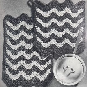 Peut inclure: Deux maniques rectangulaires au crochet présentant un motif à chevrons alternant des couleurs sombres et claires avec une petite boucle de suspension, placées à côté d'un couvercle de casserole rond, métallique et argenté.