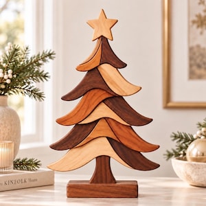 Puede incluir: Una escultura decorativa de árbol de Navidad de madera con capas alternas de madera clara, media y marrón oscura. El árbol está rematado con una estrella de cinco puntas y se asienta sobre una base rectangular, con un acabado liso y pulido.