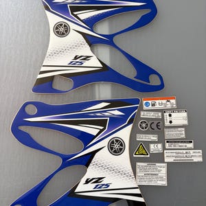 Peut inclure: Deux autocollants de radiateur de moto Yamaha YZ 125 bleus, blancs et noirs avec des motifs géométriques et le logo Yamaha. Comprend un ensemble de petits autocollants rectangulaires de sécurité, d'avertissement et d'information avec du texte.