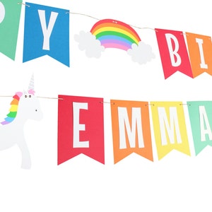 Puede incluir: Bandera arco&iacute;ris con las palabras "Happy Birthday Emma" y dos figuras de unicornio blancas con crines arco&iacute;ris.