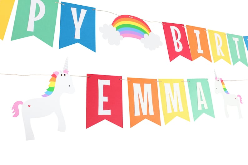 Puede incluir: Bandera arco&iacute;ris con las palabras "Happy Birthday Emma" y dos figuras de unicornio blancas con crines arco&iacute;ris.