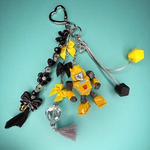 May include: A decorative keychain featuring a yellow and gray robot figure, black and yellow bows, black flower-shaped beads, a silver skull bead, a clear heart-shaped charm, a gray tassel, and two geometric beads on gray cords, all attached to a heart-shaped ring.