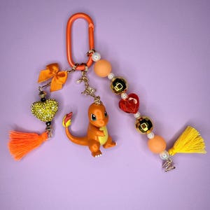 May include: A keychain featuring an orange plastic carabiner, a Charmander figure, and two beaded strands. One strand has an orange bow, a yellow rhinestone heart, and an orange tassel. The other has matte orange beads, gold metallic beads, a red heart bead, and a yellow tassel.