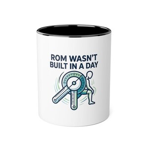 May include: A white ceramic mug with a black interior and rim. The exterior features a graphic of a person using a goniometer to measure joint range of motion, accompanied by the dark blue text that reads: ROM WASN'T BUILT IN A DAY.