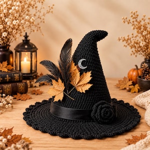 May include: A black crocheted pointed hat featuring a wide brim, a black satin ribbon band, and a crocheted rose accent. The hat is decorated with two dark feathers, two dried golden maple leaves, and a small silver crescent moon charm.