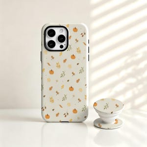 Peut inclure: Une coque de smartphone beige clair et un support de téléphone rétractable assorti, ornés d'un motif répétitif de petites citrouilles orange, de glands marron et de diverses feuilles d'automne vertes et jaunes éparpillées.