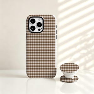 Puede incluir: Una funda para smartphone y un soporte circular plegable a juego, ambos con un patrón de cuadros vichy en marrón y blanco. La funda tiene un recorte negro para el módulo de cámara con tres lentes circulares y un flash visible.