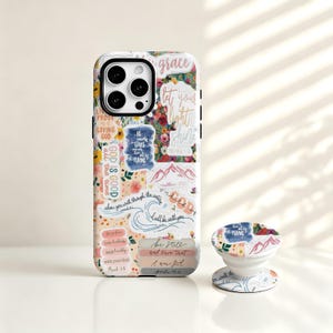 May include: A smartphone case and a matching circular phone grip featuring a collage of floral patterns, mountain illustrations, and various religious quotes including "God is good," "Be still and know that I am God," and "Let your light shine."