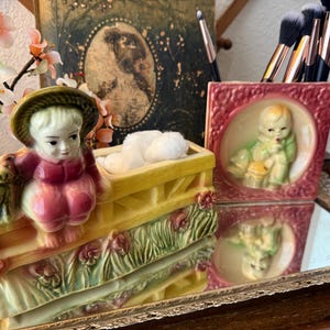 May include: A ceramic planter featuring a child in a hat sitting on a yellow fence with a small dog, alongside a square ceramic container with a relief of a child in a pink frame, both holding makeup brushes and white cotton balls.