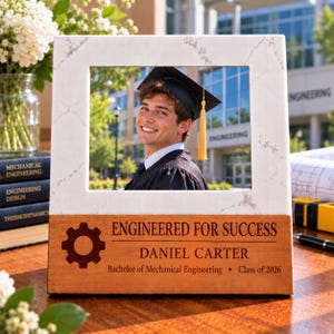 May include: A white marble-patterned picture frame with a wooden base featuring a gear icon and the text: ENGINEERED FOR SUCCESS, DANIEL CARTER, Bachelor of Mechanical Engineering, Class of 2026. The frame holds a photo of a graduate in a cap.