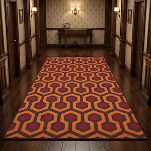 May include: A rectangular area rug featuring a repeating geometric pattern of interlocking hexagons in shades of orange, dark red, and brown. The rug is centred on a dark wooden floor within a hallway lined with patterned wallpaper and wall sconces.