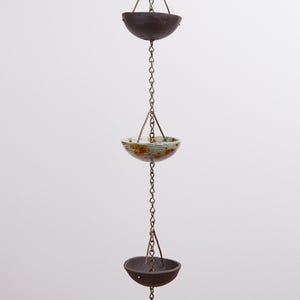 Op de afbeelding: Een regenketting met een reeks donkerbruine, mat afgewerkte keramische kommen opgehangen aan een metalen ketting. De middelste kom heeft een lichtblauw en groen glazuur met bruine spikkels, terwijl de bovenste en onderste kommen donkerbruin zijn.