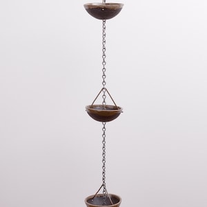 Op de afbeelding: Een verticale regenketting met een reeks donkerbruine, komvormige keramische bekers opgehangen aan een metalen schakelketting. Elke beker is met dunne metalen steunen aan de centrale ketting bevestigd, wat een herhalend patroon van hangende vaten vormt.