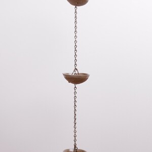 Op de afbeelding: Een verticale regenketting met een reeks bruine, komvormige keramische bekers verbonden door een donkere metalen ketting. De bekers zijn gelijkmatig verdeeld over de lengte van de ketting en elk opgehangen aan kleine metalen schakels.