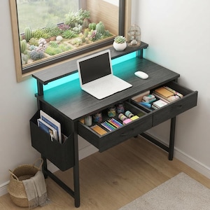 Peut inclure: Un bureau en bois noir à grain apparent doté de deux tiroirs remplis de papeterie, d'une pochette de rangement latérale et d'une étagère surélevée avec éclairage LED bleu sarcelle. Un ordinateur portable blanc et une souris sont posés dessus.
