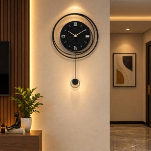 May include: A black wall clock featuring a circular face with white Roman numerals and gold hands. The clock is surrounded by two thin black concentric rings and includes a long, slender pendulum with a small circular weight at the bottom.