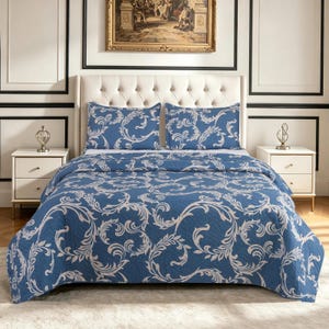 May include: A blue bedspread and two matching pillow shams featuring a white scrolling acanthus leaf pattern. The bedding set is displayed on a tufted headboard bed frame with two white nightstands on either side, each holding a silver lamp.
