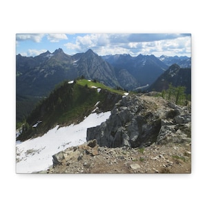 May include: A landscape view of a mountain range featuring jagged, dark grey peaks under a blue sky with white clouds. A foreground ridge shows patches of green vegetation and white snow, with rocky terrain leading toward the distant mountains.