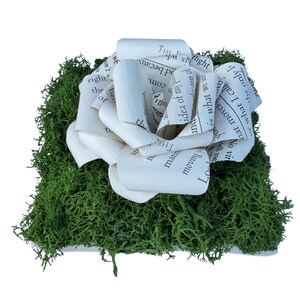 Könnte beinhalten: Eine dekorative quadratische Basis, die mit leuchtend grünem konserviertem Moos bedeckt ist, gekrönt von einer großen, mehrschichtigen Blumenskulptur aus cremefarbenen Papierseiten mit schwarzem Textdruck, einschließlich Phrasen wie the dream of my.