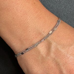 May include: A delicate double-strand silver-toned chain bracelet featuring small, flat, reflective metallic accents spaced evenly along the links. The bracelet has a fine, lightweight construction with a polished finish that catches the light.