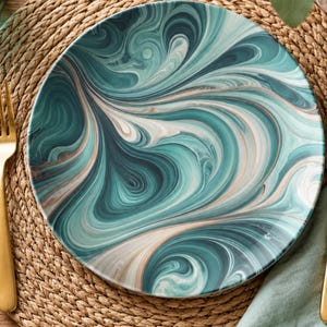 May include: A round dinner plate featuring a swirling marbled pattern in shades of teal, dark green, white, and soft beige. The plate is accompanied by a gold-toned metal fork positioned on the left and a matching gold-toned metal knife on the right.