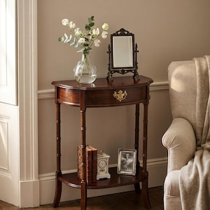 May include: A dark wood half-moon console table with a single drawer featuring a brass pull. The top holds a clear glass vase with white flowers and a small rectangular mirror. The lower shelf displays two books, a clock, and a framed photo.