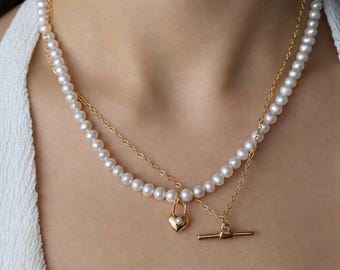 May include: A layered necklace featuring a strand of small white pearls and a gold-tone chain. The necklace includes a gold-tone heart-shaped padlock charm with a tiny pearl center and a gold-tone toggle clasp closure at the front.