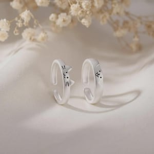 May include: Two silver-toned open rings featuring cat-inspired designs. One ring displays a cat face with pointed ears and whiskers, while the other ring shows a cat face with rounded ears and a simple nose and mouth detail, both with black markings.