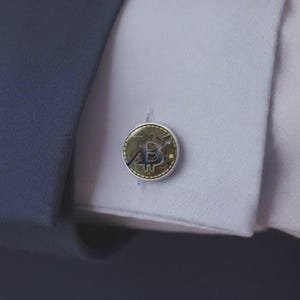 May include: A circular silver-toned cufflink featuring a gold-colored Bitcoin symbol with an upward-trending arrow graphic. The cufflink is secured through the buttonhole of a crisp white dress shirt cuff, which is partially covered by a dark blue jacket sleeve.