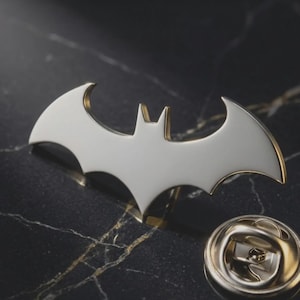 Könnte beinhalten: Eine Anstecknadel aus Metall in Form einer Fledermaus-Silhouette mit glatter weißer Emaille-Einlage und goldfarbenen Kanten. Die Nadel verfügt über einen runden Metallverschluss mit einem Schließmechanismus, der separat neben der Nadel liegt.